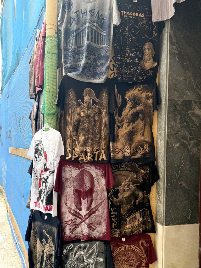 Display of various graphic t-shirts featuring themes related to ancient Greece, including images of Athens, Spartans, Poseidon, and mathematical concepts like the Pythagorean theorem.