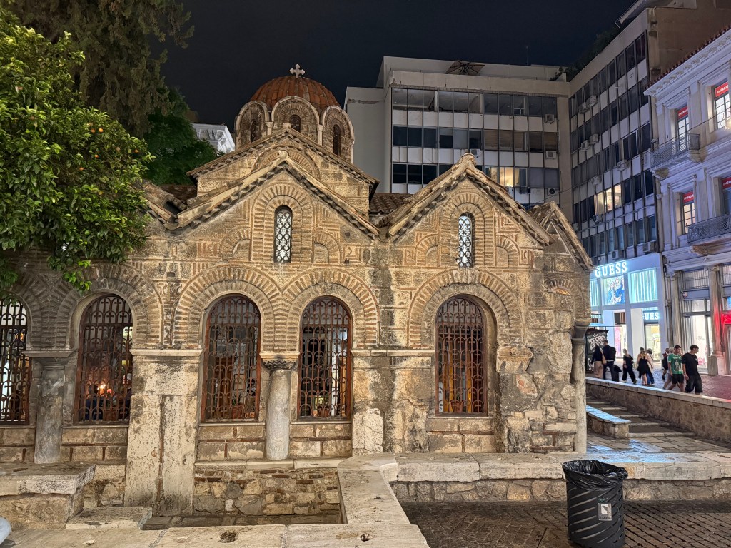 An ancient stone building with a domed roof and arched windows, surrounded by modern buildings at night.