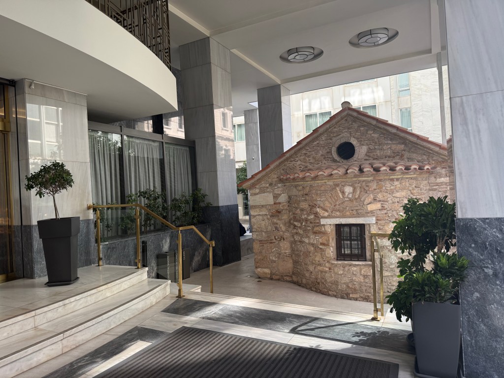 Entrance of a modern building featuring a quaint stone structure with a tiled roof, surrounded by decorative plants.