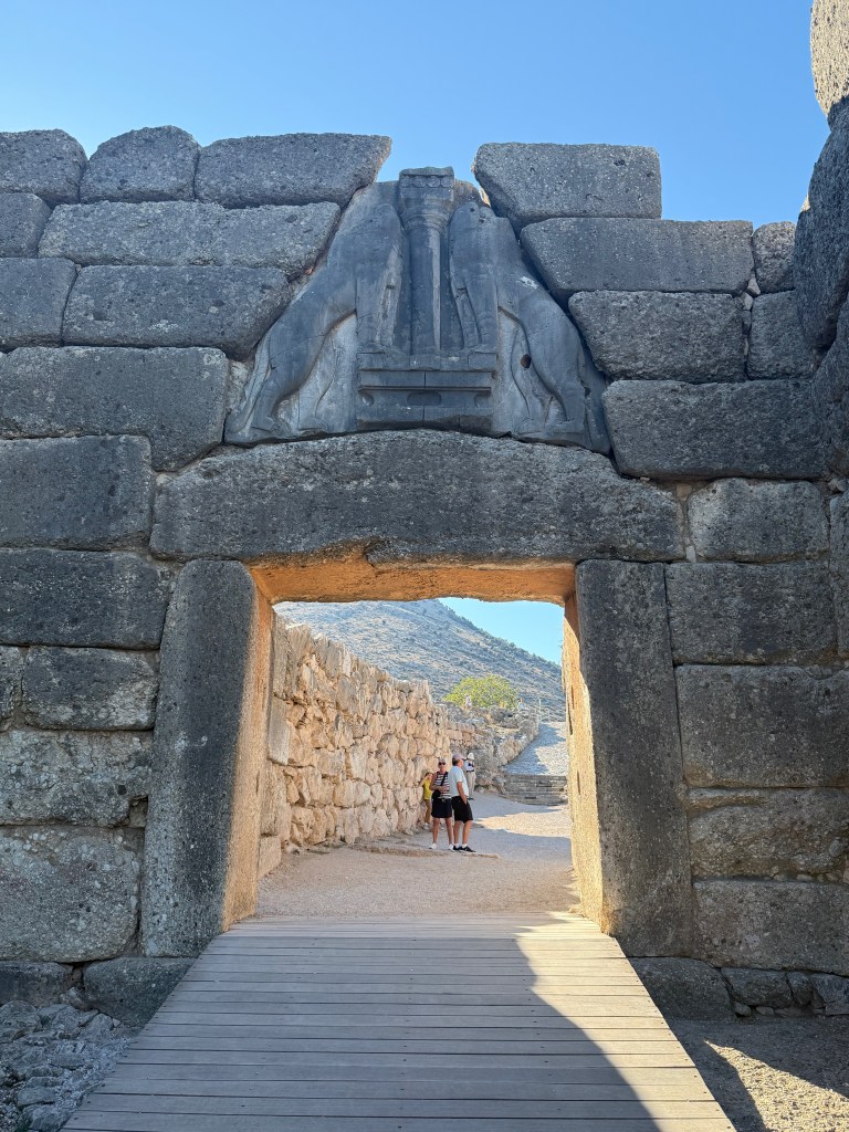 The entrance to an ancient stone structure featuring a carved lion relief above the doorway, with two people standing in front of it and mountains in the background.