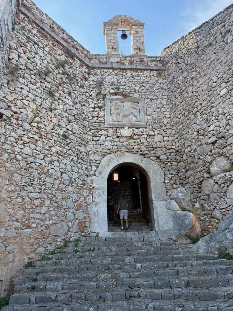An ancient stone gateway with a bell above it, leading up a staircase. The archway features a relief of a lion, surrounded by weathered stone walls.