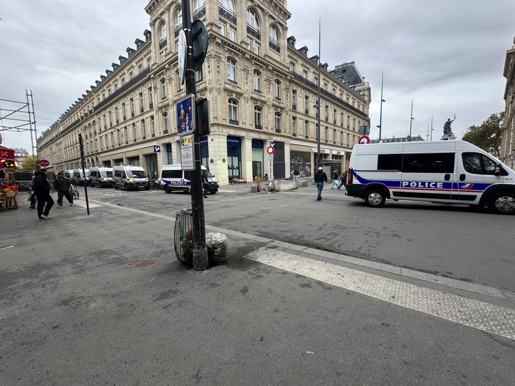 A street scene showing several police vans parked along a city street with a historic building in the background. Pedestrians are walking on the sidewalk, and a traffic signal stands on the corner.